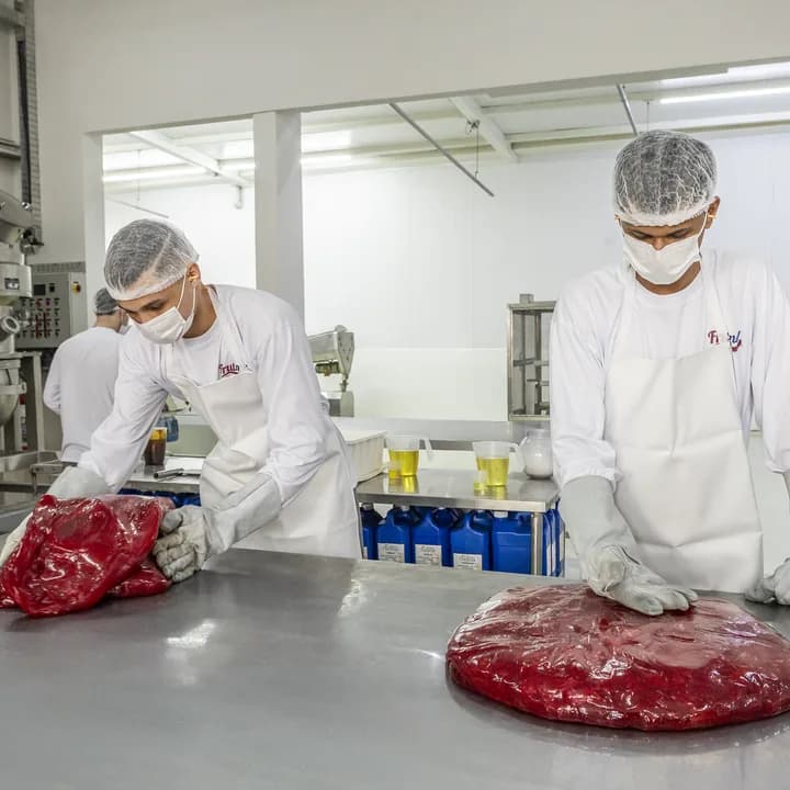 Dois profissionais de jaleco, touca e máscara trabalham a massa de bala vermelha sobre a bancada de inox