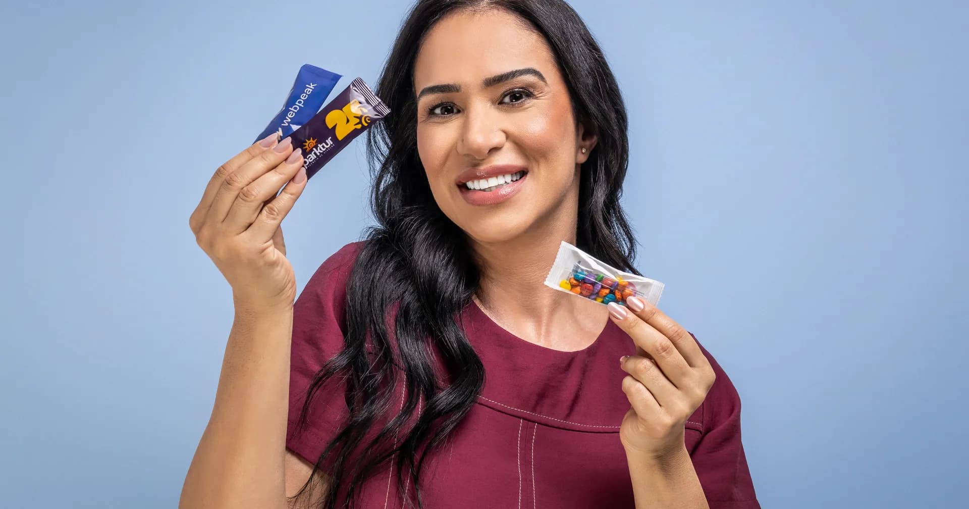 Mulher sorrindo segura duas embalagens de brinde personalizado, uma bala e um snack com a marca impressa
